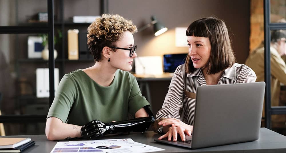 Zwei Frauen an einem Laptop. Die Eine Frau trägt eine Armprotese.